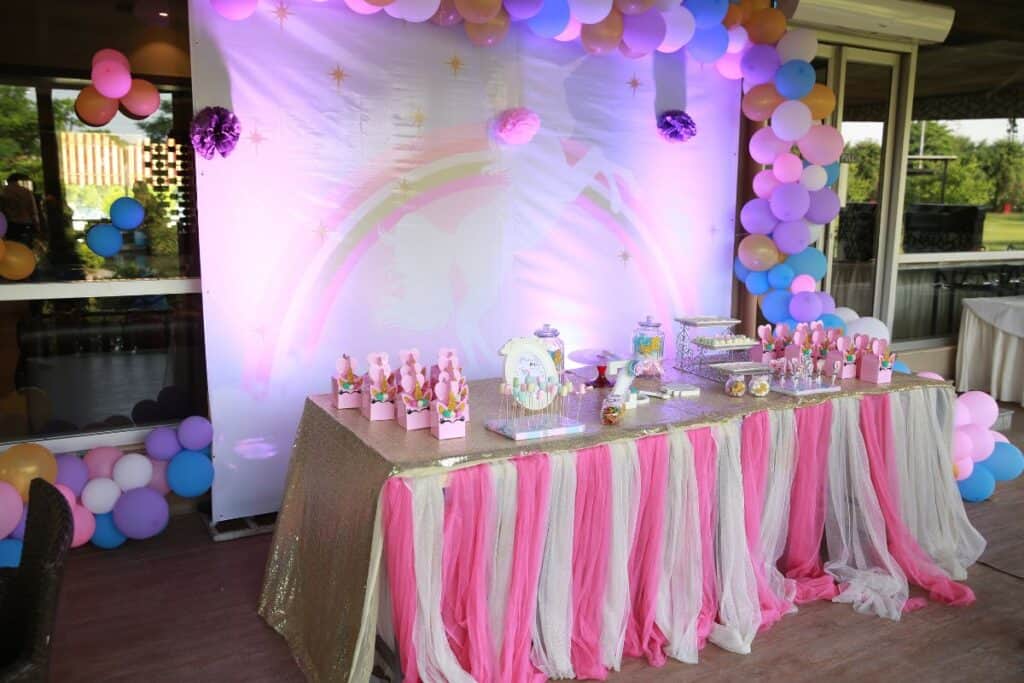 Dessert table decorated with pink and white tablecloths and a large backdrop with a balloon arch.