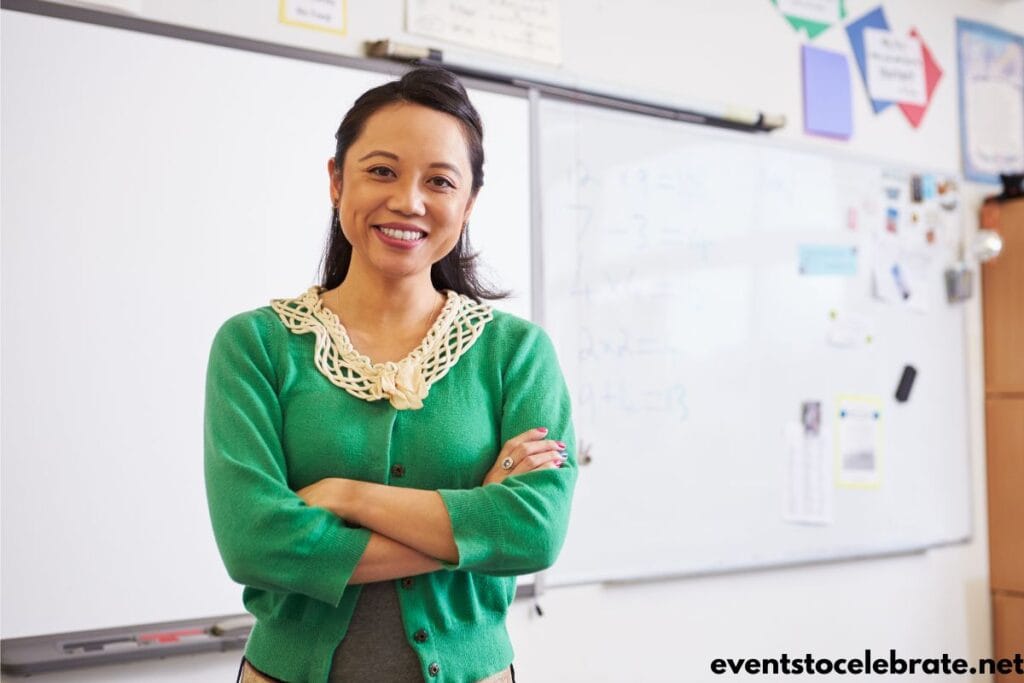A female teacher, smiling, and standing in front of her white board in her classroom.