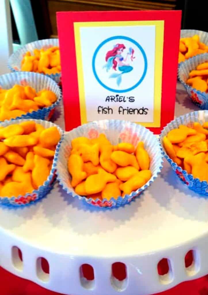 Goldfish crackers poured into cute muffin liners and put on a cake stand. The food label says, "Ariel's Fish Friends" and is perfect for this princess birthday party.